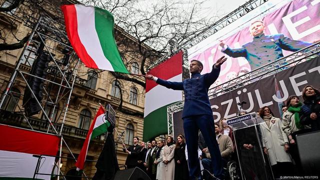 Photographed from below, Peter Magyar in a dark-blue suit with distinctive jacket buttons stands on a stage waving a Hungarian flag; his image is being projected behind him on a screen, and a few dozen people are standing at the back of the stage. | Image: Marton Manus/REUTERS