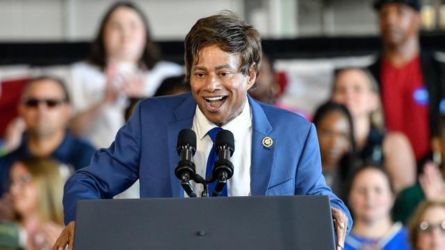 Rep. Shri Thanedar wearing a blue suit and speaking at a gray podium in front of a crowd of people. Rep. Shri Thanedar wearing a blue suit and speaking at a gray podium in front of a crowd of people.
