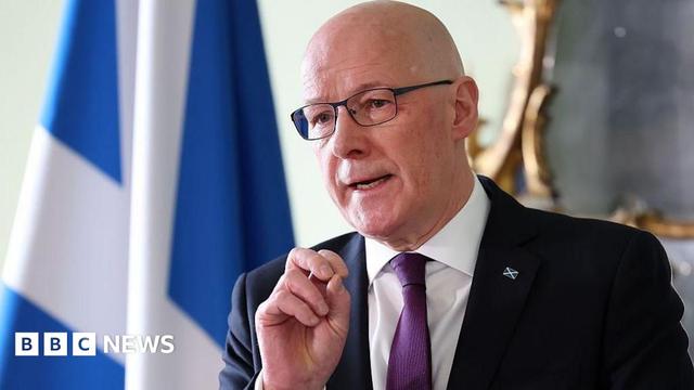 John Swinney, a blad man waring glasses, a dark suit with white shirt and purple tie, is standing in front of a Saltire flag with his right hand raised as he answers a question from an unseen group John Swinney, a blad man waring glasses, a dark suit with white shirt and purple tie, is standing in front of a Saltire flag with his right hand raised as he answers a question from an unseen group