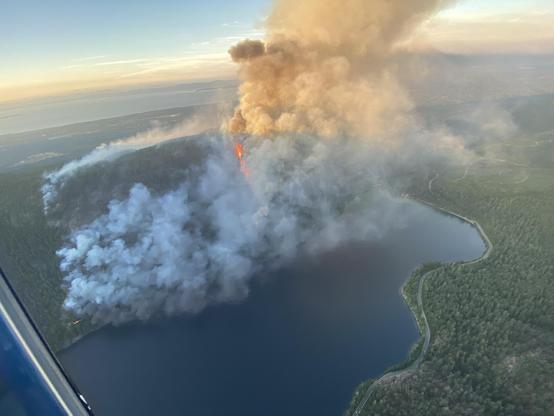 An aerial view of the fire at Wesley Ridge and Cameron lake shows a wide swath of smoke with fire reaching the top of the mountain. At the bottom left, a line of fire appears to be along the railway, possibly the trestle.