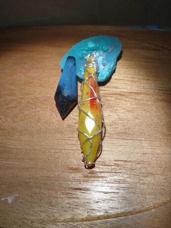 Two crystal shaped pendants, one blue and black and the other yellow, red, and orange wrapped in silver wire, both resting on a wooden tray and a blue resin geode that is propping them up.