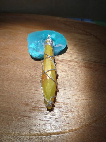 The back of a crystal shaped pendant that is yellow, red, and orange wrapped in silver wire, resting on a wooden tray and a blue resin geode that is propping it up.