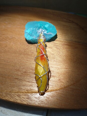 The front of a crystal shaped pendant that is yellow, red, and orange wrapped in silver wire, resting on a wooden tray and a blue resin geode that is propping it up.