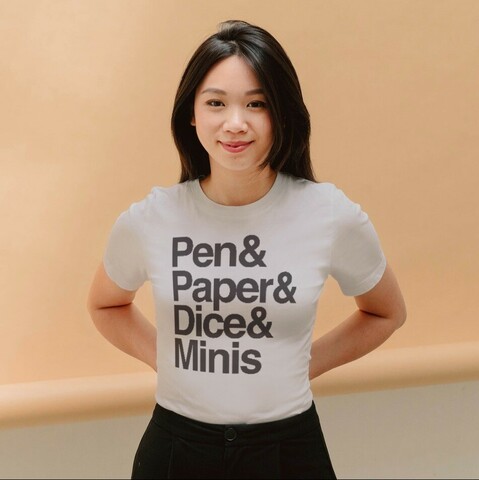 A smiling woman wearing a t-shirt that reads "Pen & Paper & Dice & Minis."