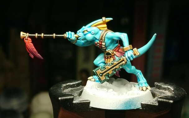 A miniature seraphon, a blue-skinned reptile humanoid. He wields a blowpipe and a jade mace, and wears gold jewellery, all decorated with  huge red-to-purple feathers.