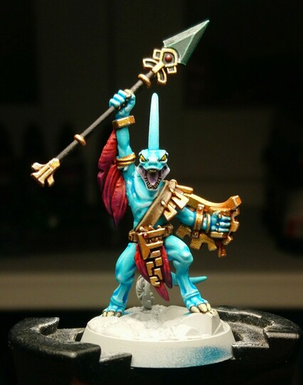 A miniature seraphon, a blue-skinned reptile humanoid. He wields a jade-tipped spear and golden shield, and wears elaborate gold jewellery decorated with  huge red-to-purple feathers.