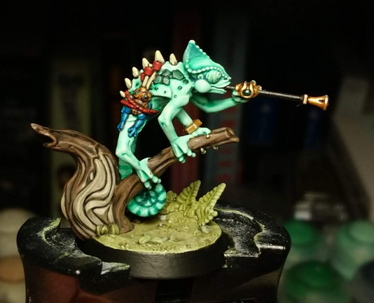 A miniature seraphon chameleon skink, a green-skinned reptile humanoid. He is perched on a branch, wielding a blowpipe. He wears gold jewellery, and has a poison arrow frog strapped to his side, used as a pouch to carry his blowpipe darts.