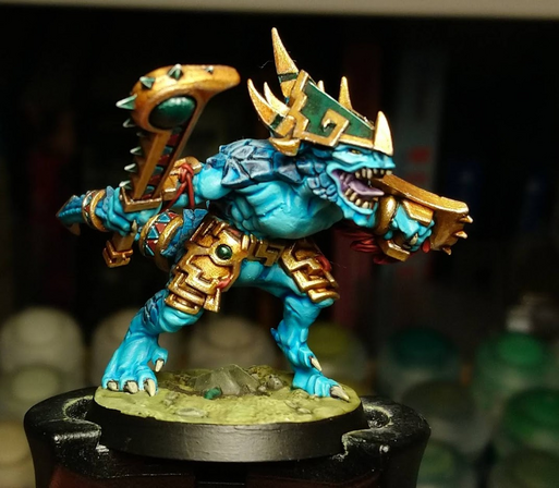 A miniature seraphon saurus, a blue-skinned reptile humanoid. He wields a gold mace inlaid with jade and other blue and red precious stones, and  a gold shield covered on one side by red scales. He wears gold jewellery, all decorated with  inlays of jade and other blue and red precious stones.