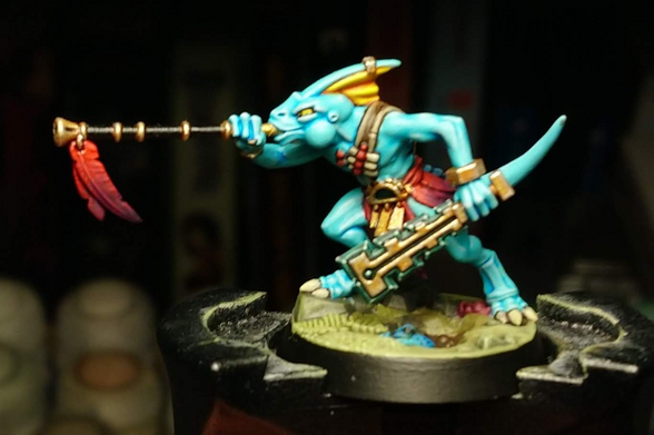 A miniature seraphon, a blue-skinned reptile humanoid. He wields a blowpipe and a jade mace, and wears gold jewellery, all decorated with  huge red-to-purple feathers. A small blue frog sits in a pool of water on his base.