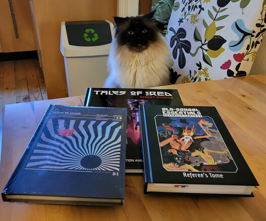 A photo of three books on a small timber table, with Jimothy the rag-doll cat sitting on a chair with a floral cloth cover at that table. He's very handsome. The books are "Cyberpunk Red Tales of the Street", "Death in Space", and "Old-School Essentials Advanced Fantasy Referee's Tome".