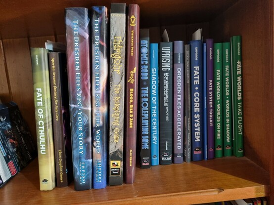 A bookshelf with the following RPG books: Fate Of Cthulhu, Aperita Arcana: Fantasy For Fate Core, The Dresden Files RPG: Your Story, The Dresden Files RPG: Our World, Ehdrigohr: The Roleplaying Game, TianXia: Blood Silk & Jade, Atomic Robo The Roleplaying Game, Shadow of teh Century, Uprising: The Dystopian Universe Roleplaying Game, Dresden Files Accelerated, Inverse World Accelerated, Fate Core System, Fate Accelerated, Fate System Toolkit, Fate Worlds: Worlds On Fire, Fate Worlds In Shadow, and Fate Worlds: Take Flight.