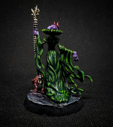 Photo of a painted miniature. A purple skinned Mind Flayer in green robes heft a spine-bone staff, accompanied by an Intellect Devourer. This photo is from the rear, revealing trailing tendrils of cloth.