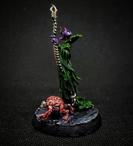 Photo of a painted miniature. A purple skinned Mind Flayer in green robes heft a spine-bone staff, accompanied by an Intellect Devourer. This photo is from the left side, focusing on the staff & Intellect Devourer. 