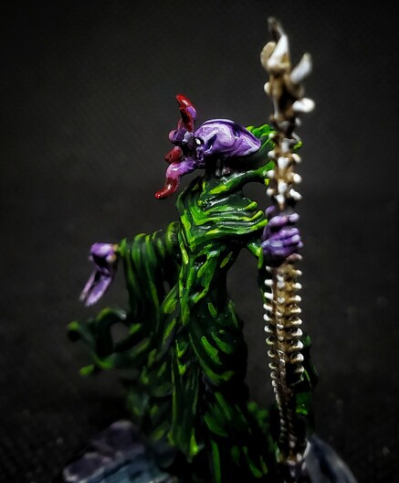 Photo of a painted miniature. A purple skinned Mind Flayer in green robes heft a spine-bone staff, accompanied by an Intellect Devourer. This angle focuses on the facial features.