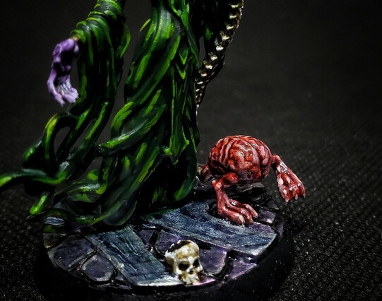Photo of a painted miniature. A purple skinned Mind Flayer in green robes heft a spine-bone staff, accompanied by an Intellect Devourer. This angle focuses on the Intellect Devourer. 