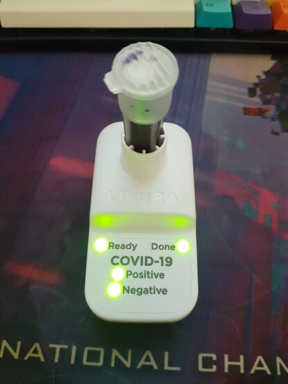 an at home COVID test with a plastic body and a vial inserted on top. the lights are labelled ready, done, positive, and negative - all of the lights are lit green