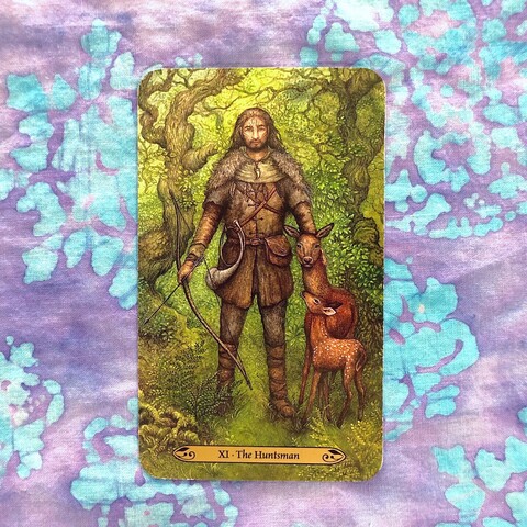 A rectangular card with a painting of a ling haired, bearded man standing in a green woods, twisted and gnarled trees visible in the background. His face is tattooed/painted and he is wearing hunting garb including a fur cloak, a feather trimmed hood and a necklace with a large tooth. There is a pack hanging from one side of his belt and a hunting horn on the other. In his right hand he hold a knocked bow at rest, his left hand rests gently on the neck of a doe who is looking warily at the viewer while her fawn stands in front of he looking up at the huntsman.