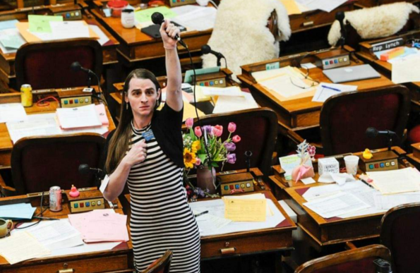 Photo of a very alone-looking Montana state rep Zooey Zephyr standing amidst a sea of desks, holding a microphone over her head, with a look of concern on her face.