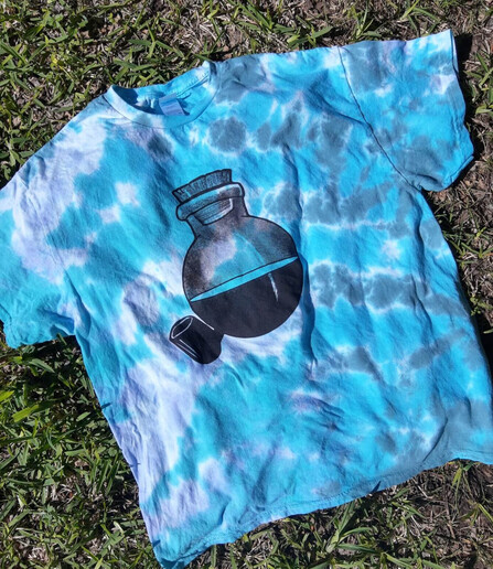 A tie dye shirt in a dreamy cloudy blue, grey and white with hints of subtle greenish tones. The image on the garment is printed with black ink; a potion bottle with a pipe in the linework and halftone style of Bob Dobbs from the Church of the Subgenius