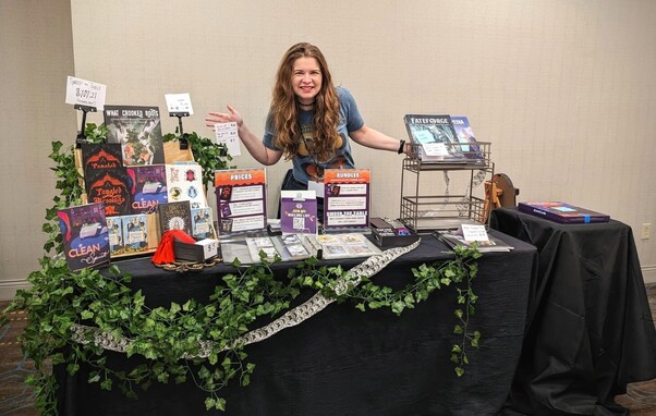 Cassi Mothwin standing behind a table showing her games. The games are Clean Spirit, Tangled Blessings, and What Crooked Roots. There are other items like tarot cards and used books pictured as welll. 