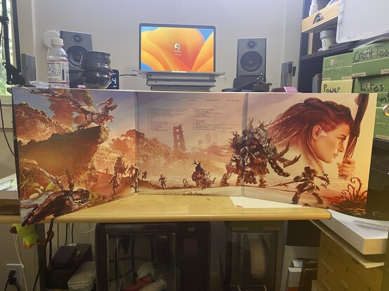 Fully open trifold album with a painted illustration spanning all three panels of Aloy fighting dinosaur robots. 