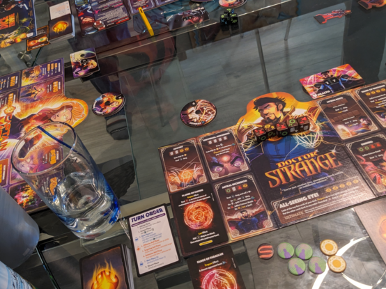 Photo of a few comic-art character mats at one end of a table (the other players' mats are out of view of the photo). The visible characters are Doctor Strange, Captain Marvel, and Black Widow. There's cards and dice and tokens scattered about.
