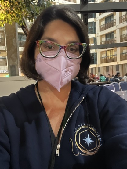 A picture of a woman with light skin and a dark brown Bob haircut. She is wearing rainbow glasses and a pink KN95 face mask. 