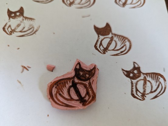 A linocut stamp showing a kind of goblin like cat holding a shield and sword. It sits on a piece of paper with several test stamps, some from when it was in progress 