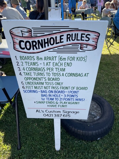 A signpost that reads:

“Cornhole Rules

1. Boards 8m apart (6m for kids)
2. 2 teams—1 at each end
3. 4 cornbags per team
4. Take turns to toss a cornbag at opponent’s board
5. Underarm toss only
6. Feet must not pass front of board

Scoring:
Bag on board = 1 point
Bag in hole = 3 points
1st team to 21 points wins!

Swap ends & play again!!
Have fun!!!

Al’s Custom Signage
0421 387 615”