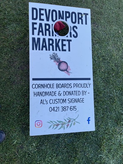 A cornhole board—roughly 4 feet long and two feet wide, raised up at the top end on 6-inch legs.

The top half, pierced with a 4-inch hole approximately 4 inches from the top edge, advertises the Devonport Farmers Market; the bottom half reads:

“Cornhole boards proudly handmade & donated by Al’s Custom Signage 0421 387 615”