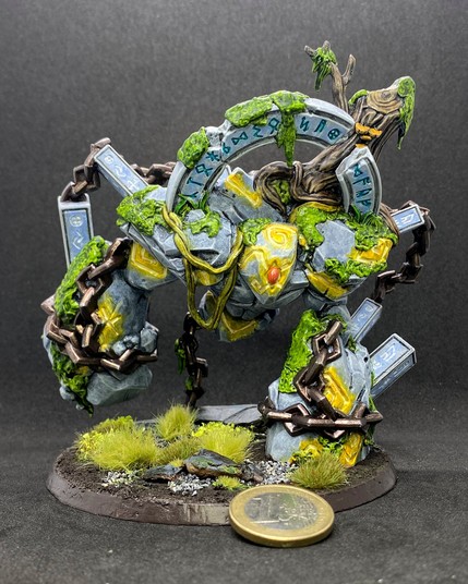 Painted miniature of an animated stone construct, hulking stompily on its 2...legs? (they come out where the arms would be on the torso). It has a runed arch over its head and pillars coming out of its legs and back with rune engravings, connected to chains which are wrapped around its legs. It is mossy and also has a tree stump and vines on it. Both the carved runes and the magical symbols on its body have a glowing effect.