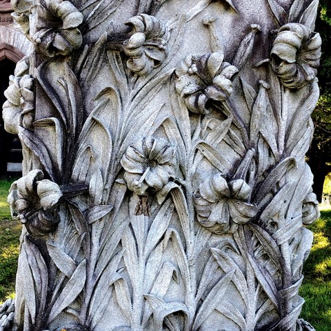 Photo of part of an older grave marker: an apparent pillar of pale stone covered in lilies in full bloom, the flowers are carved out in 3D and the crevices and parts have turned dark with age and weather highlighting the flowers and leaves that much more and making them appear even more dramatic.
In the center of the pillar “hiding” under one of the flowers is a large brown moth (although it appears small in comparison to the flowers)