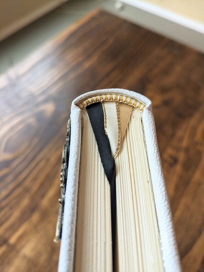The top of a journal showing a hand made endband made of gold and white silk. The journal has 3 ribbon bookmarks in black, white, and gold.