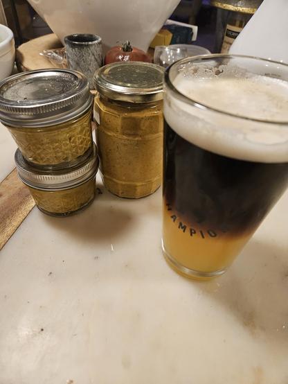 A pint glass of  half guinness, half winter cider, and three jars of my homemade blazing hot mustard on a light countertop.