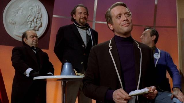 A still from the TV show, The Prisoner, with Patrick McGoohan's character, Number 6, standing in front of some heavies from The Village.