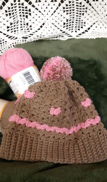 A tuque sits on a green background. The tuque is a light brown color and pink. There are pink hearts in a row along the second row, and intermittent pink hearts crochet's around it.  There is a brown ribbing for around the temples. There is a pink pompom at the top with little bits of brown speckled in it. Two skeins of yarn sit to the left side of the hat. One is brown and the other is pink. And, in the background is my Great Great Gramma's table cloth crocheted in white hanging down as a backgdrop.