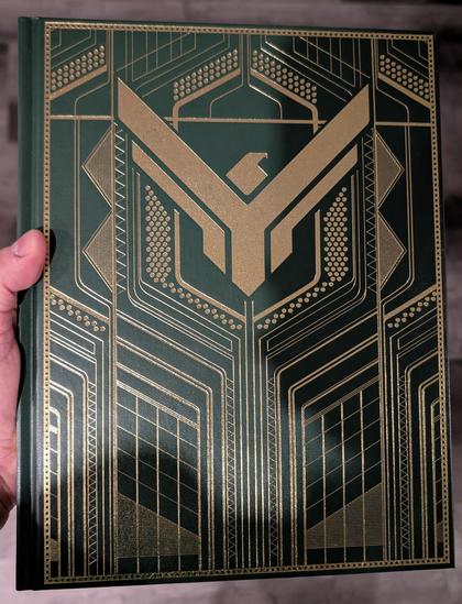 A photo of a hand holding Dune: Adventures in the Imperium RPG collector's edition (House Atreides variant) core book.