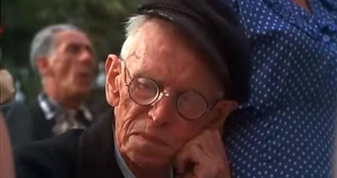 A seated elderly German man wearily rests his head on his hand as a crowd stands and sings. Image is from the 1972 movie Caberet. Context is a crowd fervently joining in with a nationalistic song being sung by a member of the Hitler Youth