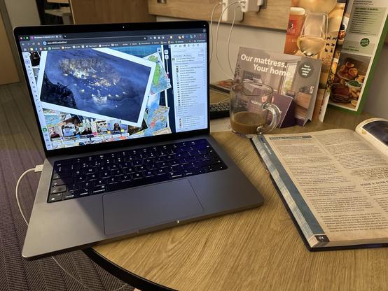 A MacBook Pro on a round hotel table with the Roll20 VTT showing dominated by a map. To the right are the hotel brochures for mattresses, drinks and more, a half drink cup of coffee and the campaign book. 