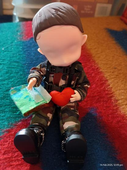 OB11 / Nendoroid scale BJD figure (6 inch doll) sitting on a blue/red/green striped fabric, and  holding a green camo satchel and a tiny red fabric heart.

Doll has short brown hair and facial indentations but no actual features. Doll is also wearing a dark camo jumpsuit with black boots.
