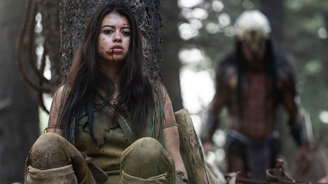 A young Comanche woman crouching hidden behind a tree.
In the background, slightly out of focus, a Predator alien is looming.