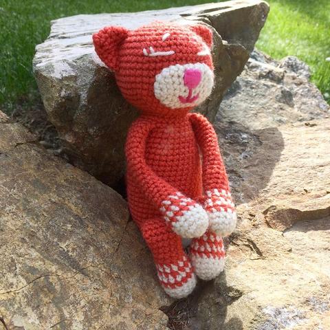 An orange crochet amineko cat with a white muzzle and white paws sits on some rocks in the sun, its legs pulled up and its paws resting on is knees as it leans back against a large rock and gazes into the distance in silent reverie