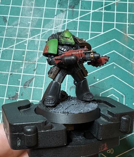A Salamander Space Maarine Adherent. His helmet and right pauldron are green. His armor is black. He is holding a red combiflamer with a chain bayonet. His helmet is shaped like a dragon or lizard. Flames are visible on the right pauldron. His left pauldron, barely visible, is a dark gold. 
