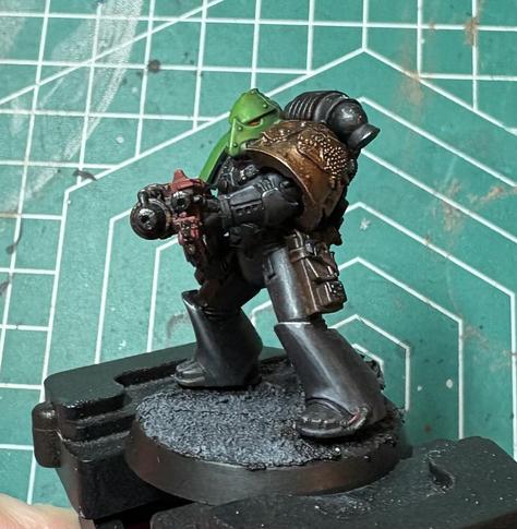 A Salamanders Adherent. His armor is black. His left pauldron is a dark gold, with a raised Salamander / Dragon sigil. His helmet is green, and shaped like a dragon. His combiflamer is red. 