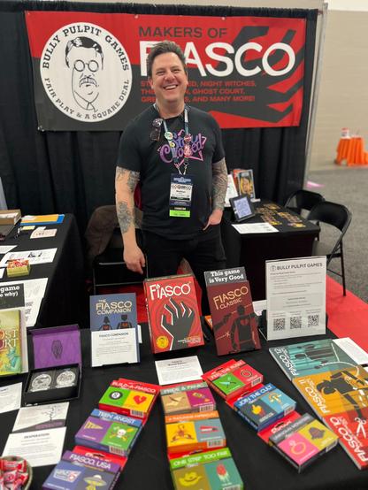 Nathan stands behind an array of Fiasco Decks and Box sets. He is wearing a Star Crossed t-shirt and behind him is a wide banner advertizing Bully Pulpit Games Makers of Fiasco.