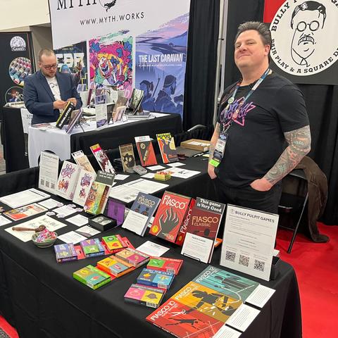 A photograph of Nathan standing behind an array of Fiasco Decks and Box sets. He is wearing a Star Crossed t-shirt. Behind him you can see the Bully Pulpit Games logo and to his right, a man stands at the Myth.Works booth. 