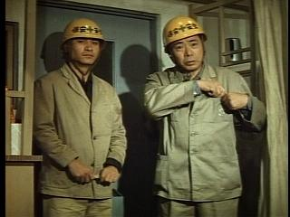 Two men in a Godzilla movie wearing yellow emergency helmets and tan work suits.