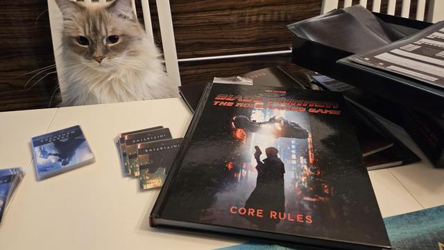 Table that has Blade Runner RPG core rulebook and cards from the Blade Runner RPG Starter Set. Siberian cat is watching the table curiously.