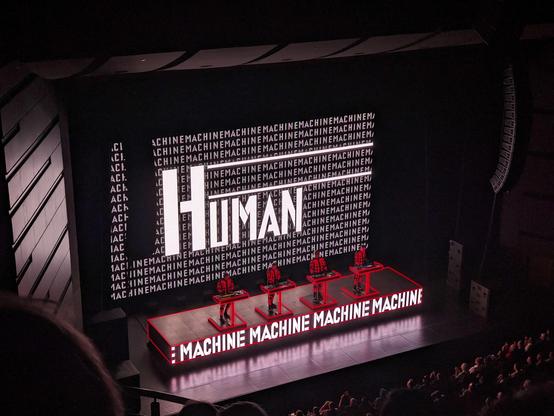 Kraftwerk members on stage performing the song "Die Mensch Maschine" on consoles. The screen behind them displays the word "Human" and the platform they're on displays "machine".