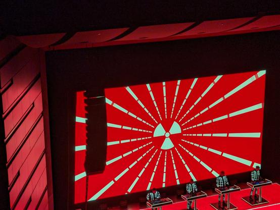 Kraftwerk members on stage performing the song "Radioactivity" on consoles. The screen behind them displays a dark orange and yellow radioactive warning sign that is radiating orange rays menacingly.